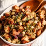 A close-up of a festive stuffing recipe in a baking dish, featuring toasted bread cubes, cranberries, celery, and herbs.