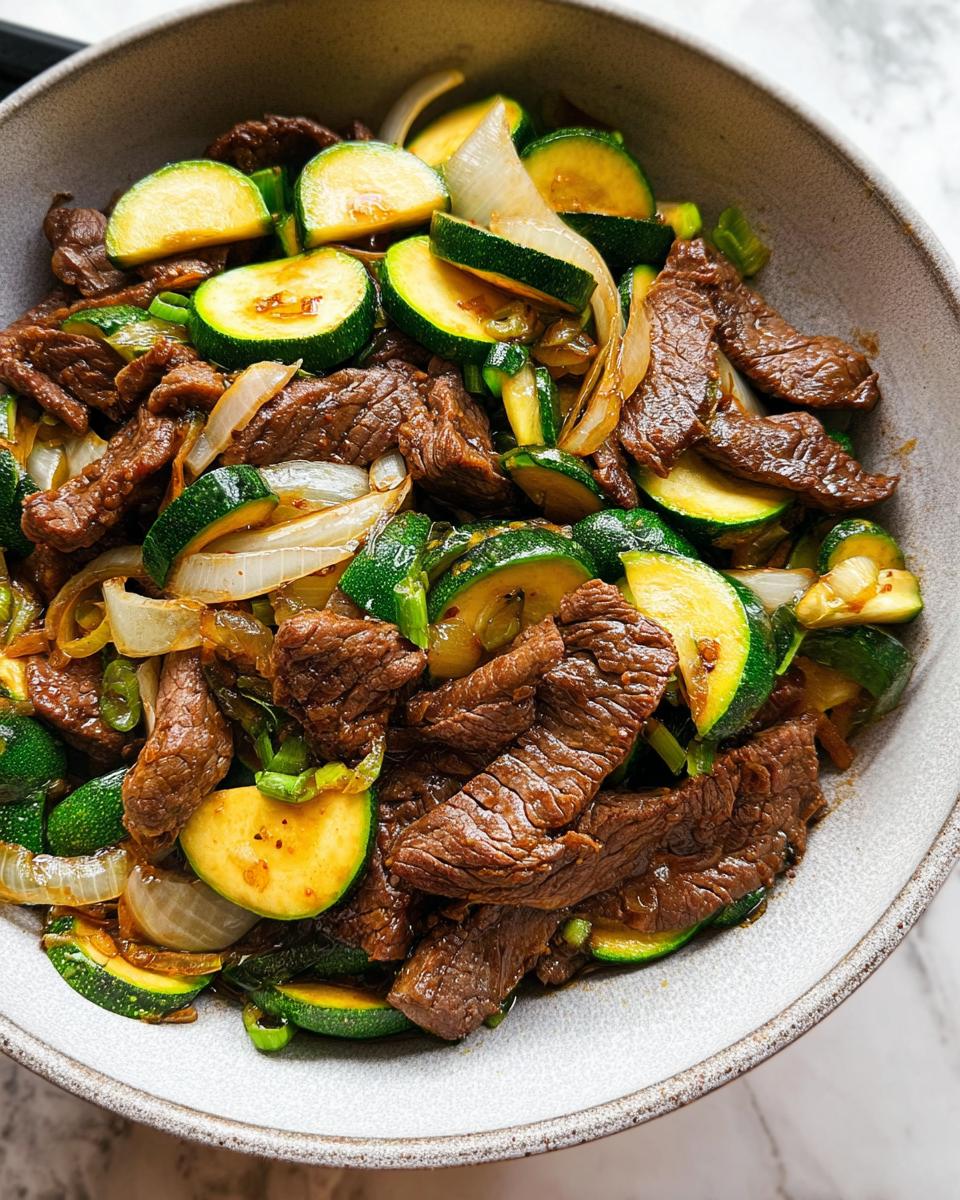 A close-up overhead view of a Cozy Grilled Steak Bowl with Zucchini, featuring sliced steak, zucchini rounds, and onions.