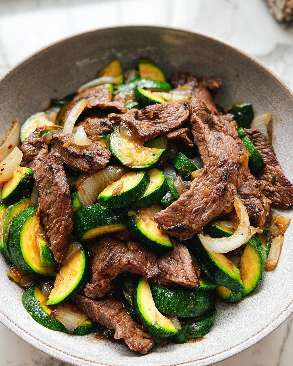 A close-up of a Cozy Grilled Steak Bowl with Zucchini, featuring sliced grilled steak, zucchini rounds, and onions in a savory sauce.