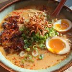 A close-up of a bowl of Creamy Garlic Chicken Ramen, featuring sliced chicken, soft-boiled eggs, and green onions.