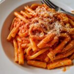 A close-up of a white bowl filled with creamy tomato garlic pasta, topped with grated cheese.