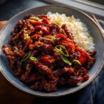 A close-up of a bowl of Crispy Chilli Beef Rice, featuring glossy beef strips and white rice, garnished with green onions.