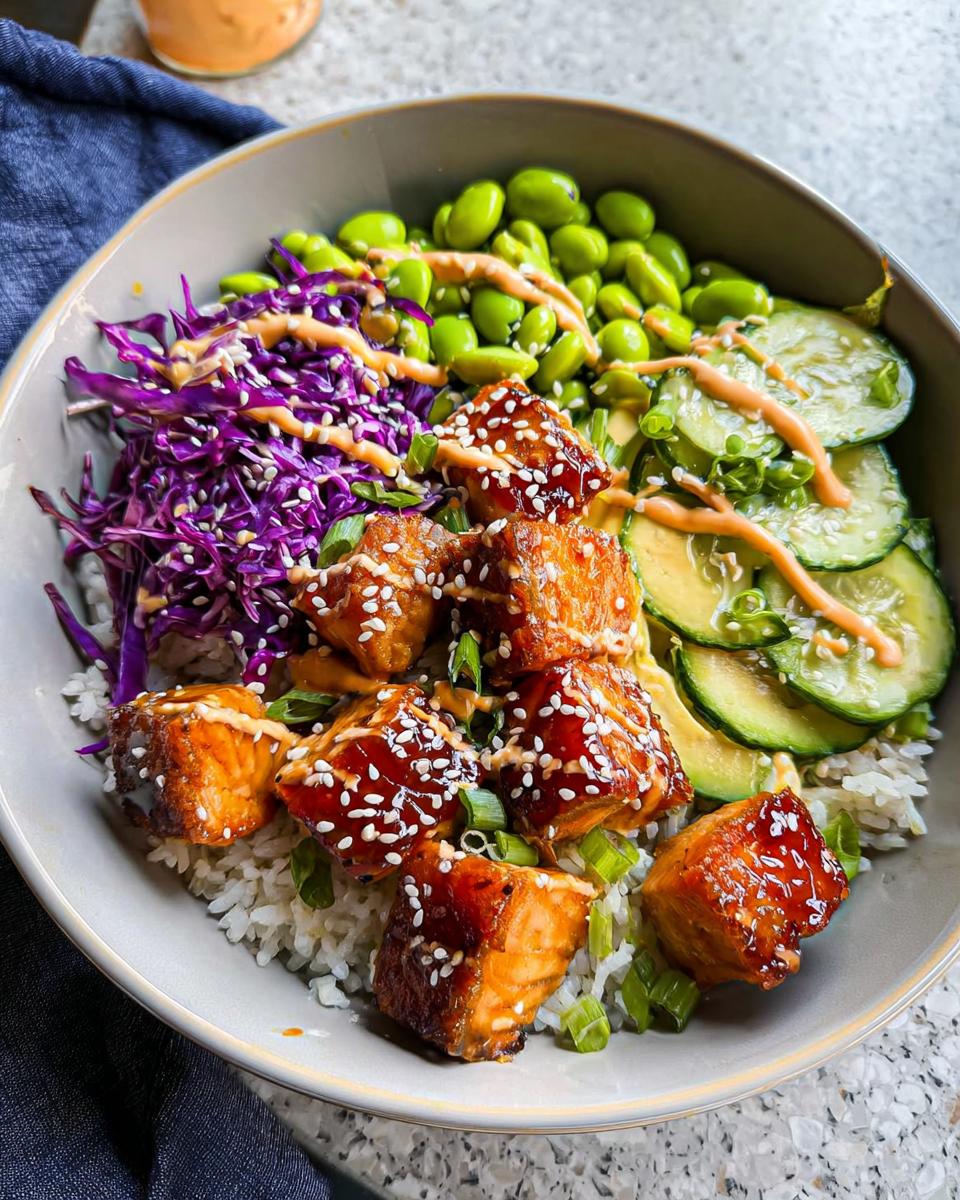 A close-up of a Crispy Salmon and Rice Bowl, featuring glazed salmon cubes, white rice, edamame, sliced cucumber, and shredded red cabbage, drizzled with sauce.