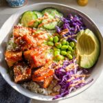 A vibrant Crispy Salmon and Rice Bowl featuring glazed salmon cubes, rice, edamame, avocado, cucumber, and red cabbage, drizzled with sauce.