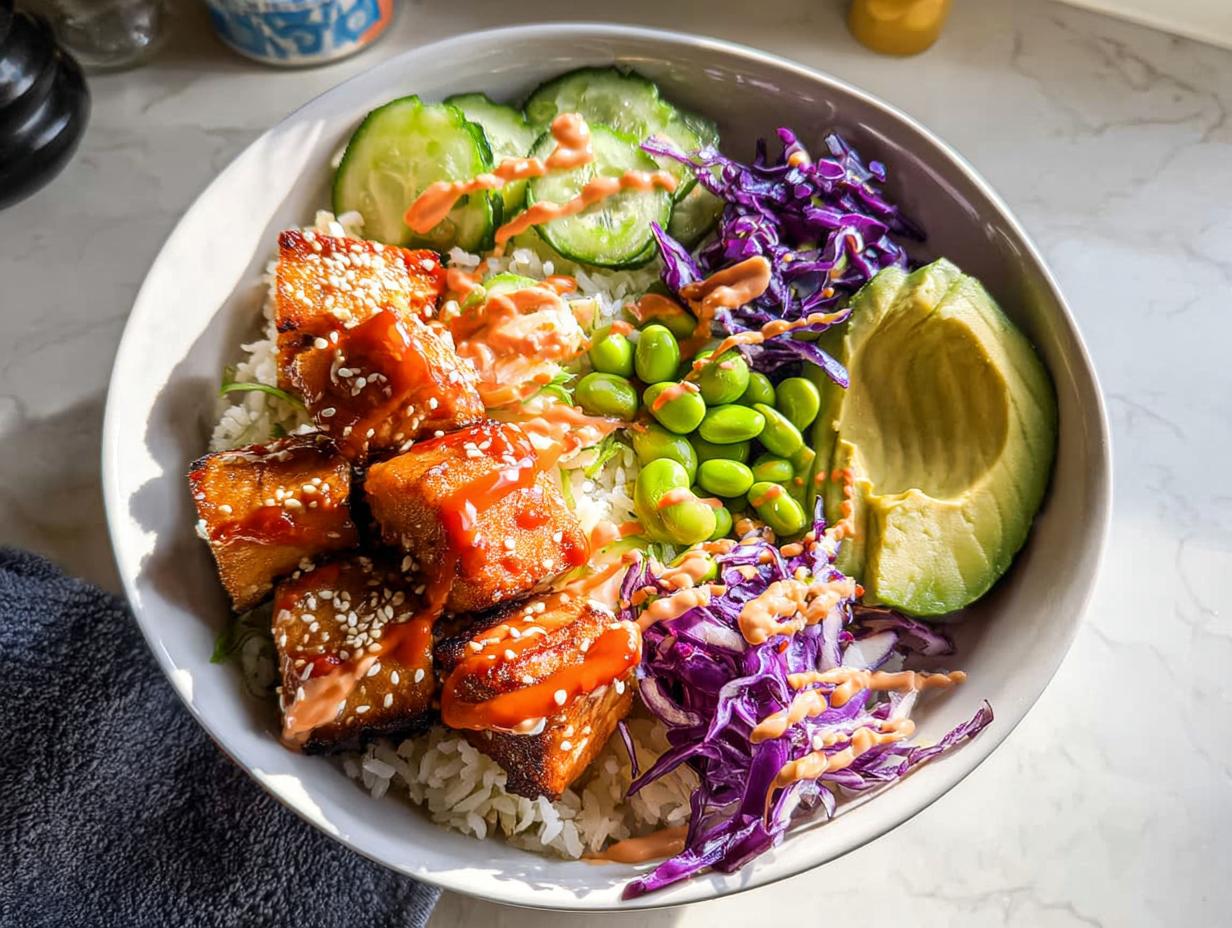 A vibrant Crispy Salmon and Rice Bowl featuring glazed salmon cubes, rice, edamame, avocado, cucumber, and red cabbage, drizzled with sauce.