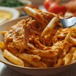 A fork lifts a portion of Crock Pot Creamy Cajun Chicken Pasta, showing tender chicken and penne pasta coated in a rich, orange sauce.