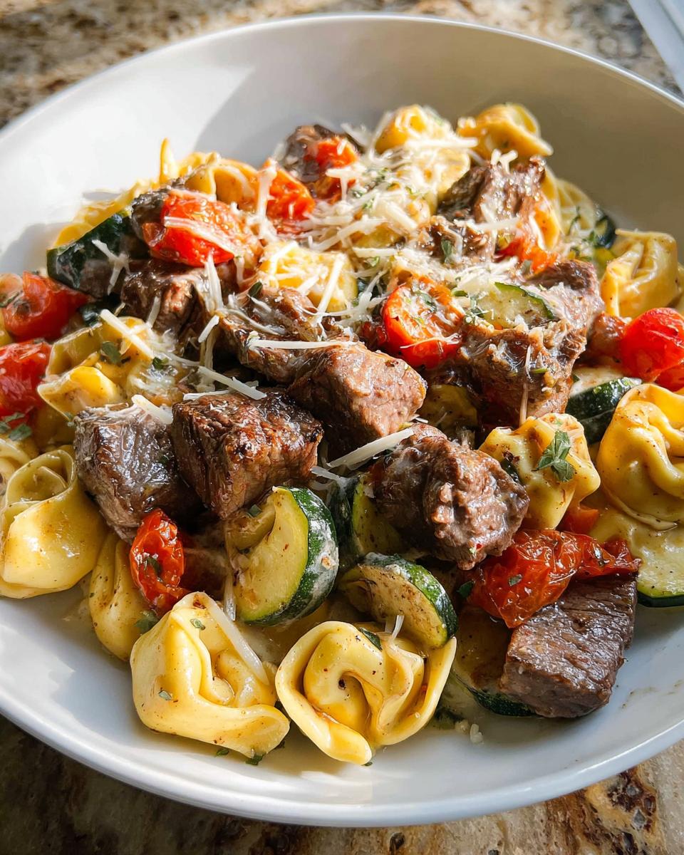 A close-up of a white bowl filled with Garlic Steak Tortellini, featuring tender steak pieces, tortellini pasta, zucchini slices, and cherry tomatoes.
