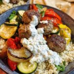 A delicious Greek Meatball Bowl Recipe featuring seasoned meatballs, grilled vegetables, and fluffy couscous, topped with a creamy sauce.