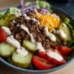 A delicious High-Protein Cheeseburger Bowl filled with seasoned ground beef, shredded cheese, pickles, tomatoes, and lettuce, drizzled with sauce.
