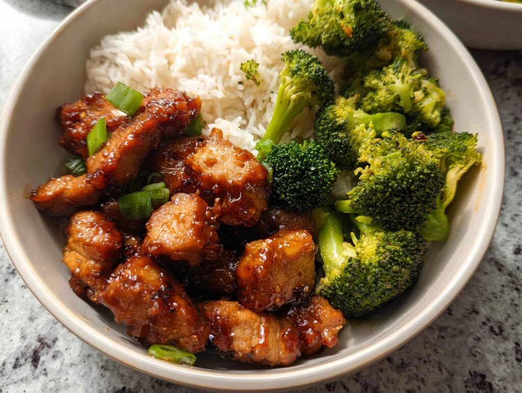 A delicious Honey Garlic Pork Rice Bowl featuring tender pork pieces, fluffy white rice, and vibrant steamed broccoli florets, garnished with green onions.