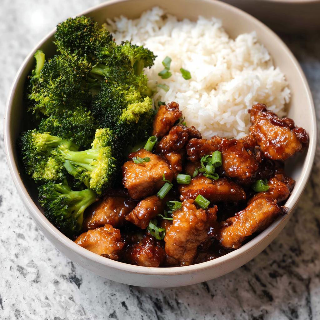 A close-up of a Honey Garlic Pork Rice Bowl with tender pork pieces, fluffy white rice, and steamed broccoli florets, garnished with green onions.