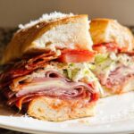 A hearty Italian Grinder Salad Sandwich piled high with meats, cheese, lettuce, and tomato on a white plate.