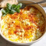 A close-up of baked feta eggs in a casserole dish, topped with paprika and fresh mint, served with pita bread.