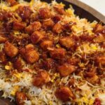 A close-up of a wooden bowl filled with fluffy rice topped with glazed BBQ chicken pieces.