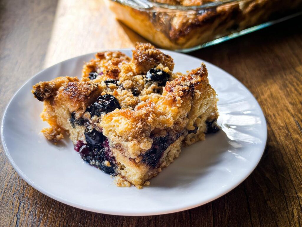 A slice of the Best Blueberry French Toast Casserole on a white plate, showing baked bread, blueberries, and a crumb topping.