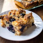 A slice of the Best Blueberry French Toast Casserole on a white plate, showing baked bread, blueberries, and a crumb topping.