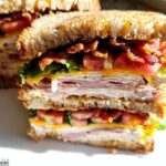 A tall, stacked Breakfast Club Sandwich with layers of turkey, ham, bacon, cheese, lettuce, and tomato on toasted bread.