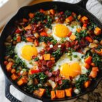 A cast iron skillet filled with a delicious BREAKFAST SWEET POTATO hash, topped with sunny-side-up eggs and crispy bacon.