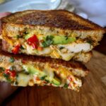 A close-up of a grilled CHICKEN AVOCADO MELT SANDWICH, showing melted cheese, avocado slices, and chopped red peppers.