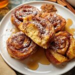 A plate of golden-brown Cinnamon Roll French Toast Bites dusted with powdered sugar and drizzled with syrup.