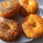 Four golden-brown cottage cheese flagels on a white plate, two topped with black sesame seeds.