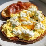 Close-up of Creamy Ricotta Egg Toast topped with scrambled eggs, ricotta cheese, and fresh herbs, with prosciutto on the side.
