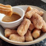 A hand dips a fluffy air fryer churro bite into a caramel sauce, with a bowl full of churro bites in the background.