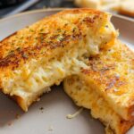 A close-up of a perfectly grilled Garlic Bread Grilled Cheese sandwich, oozing with melted cheese and sprinkled with herbs.