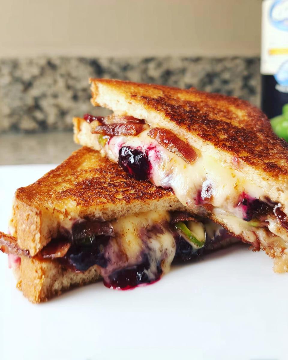 A close-up of a gourmet blueberry grilled cheese sandwich, cut in half, showing melted cheese, blueberries, and bacon.