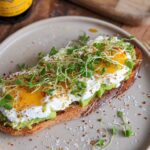 A delicious high-protein breakfast toast topped with mashed avocado, cottage cheese, a runny egg, and microgreens.