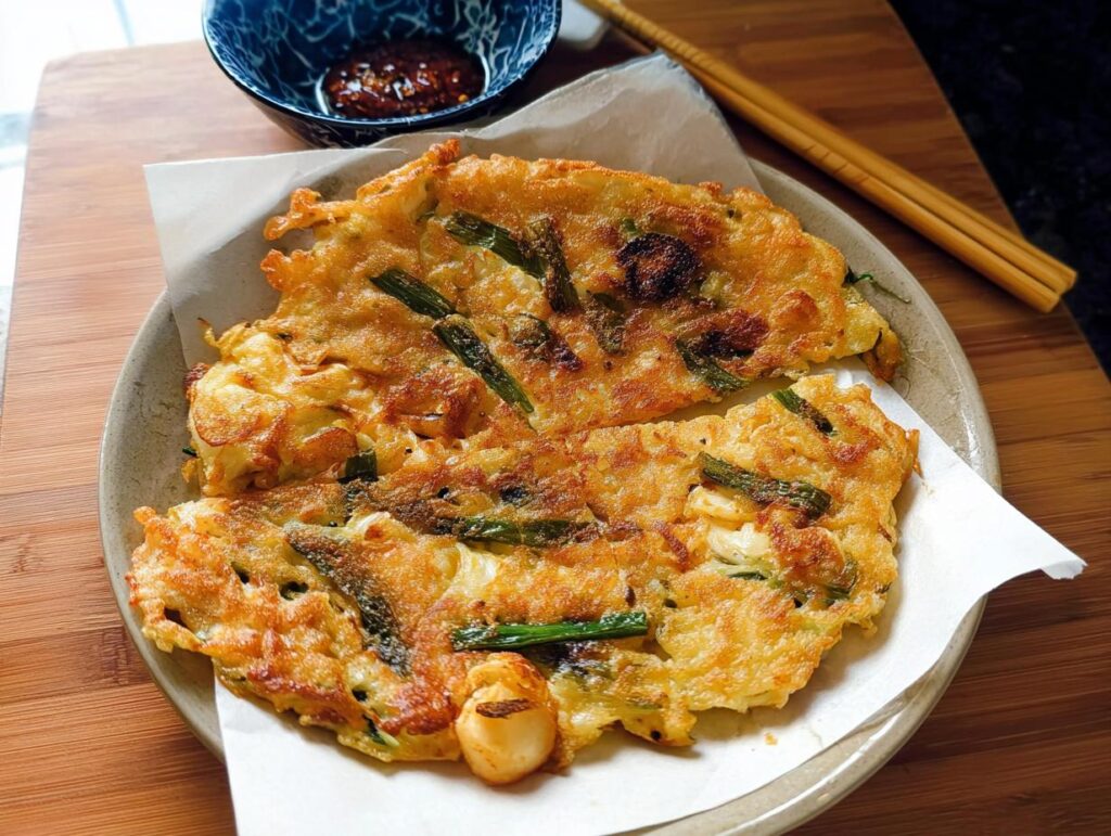 A close-up of golden-brown Korean Pancakes (Pajeon) served on a plate with a side of dipping sauce.