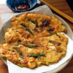 A close-up of golden-brown Korean Pancakes (Pajeon) served on a plate with a side of dipping sauce.