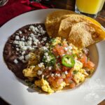 A plate of scrambled Mexican eggs topped with pico de gallo, jalapeños, and queso fresco, served with refried beans and tortilla chips.