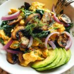 A close-up of Mushroom Spinach Scrambled Eggs topped with sliced red onion, fresh dill, and avocado.
