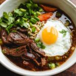 A comforting bowl of Slow Cooker Beef Ramen Noodles topped with a fried egg, shredded beef, and fresh herbs.