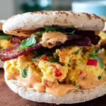Close-up of a Spicy Chipotle Egg Breakfast Sandwich with scrambled eggs, bacon, and chipotle sauce on an English muffin.