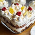 A close-up of a layered Chocolade Lasagne dessert in a glass dish, topped with meringue, raspberries, and lemon slices.