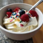 A bowl of easy frozen Greek yogurt topped with fresh raspberries, blueberries, sprinkles, and a drizzle of honey.
