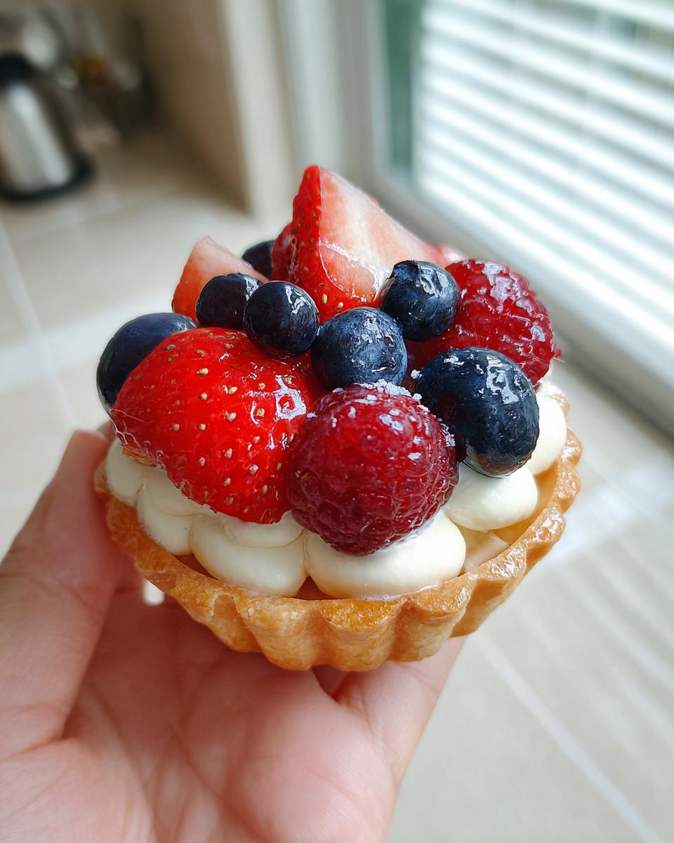 A hand holding a mini tartlet filled with cream and topped with fresh strawberries, blueberries, and raspberries, perfect for Fourth of July desserts.