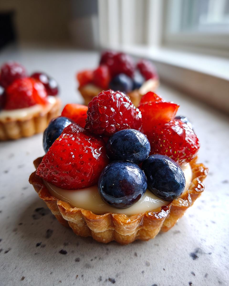 Close-up of a mini tartlet filled with cream and topped with fresh strawberries, blueberries, and raspberries for Fourth of July desserts.