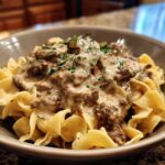 A close-up of a bowl of creamy beef stroganoff served over egg noodles, garnished with parsley. A delicious ground beef recipe.