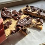A hand holding a slice of healthy cookie dough bark topped with chocolate chips and sea salt.