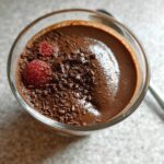 A decadent serving of High Protein Chocolate Pudding topped with chocolate shavings and fresh raspberries in a glass.