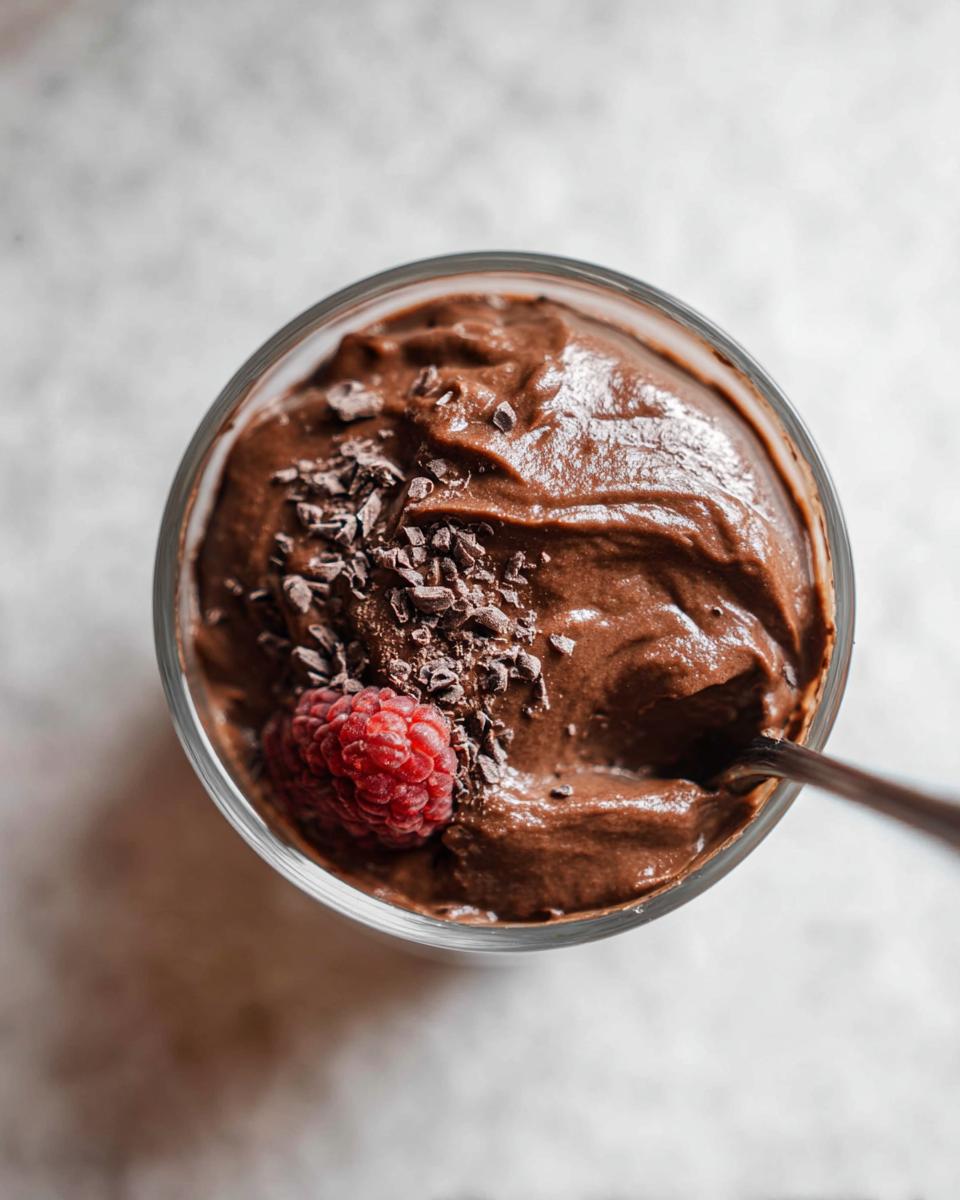 A close-up of a glass filled with rich High Protein Chocolate Pudding, topped with chocolate shavings and a fresh raspberry.