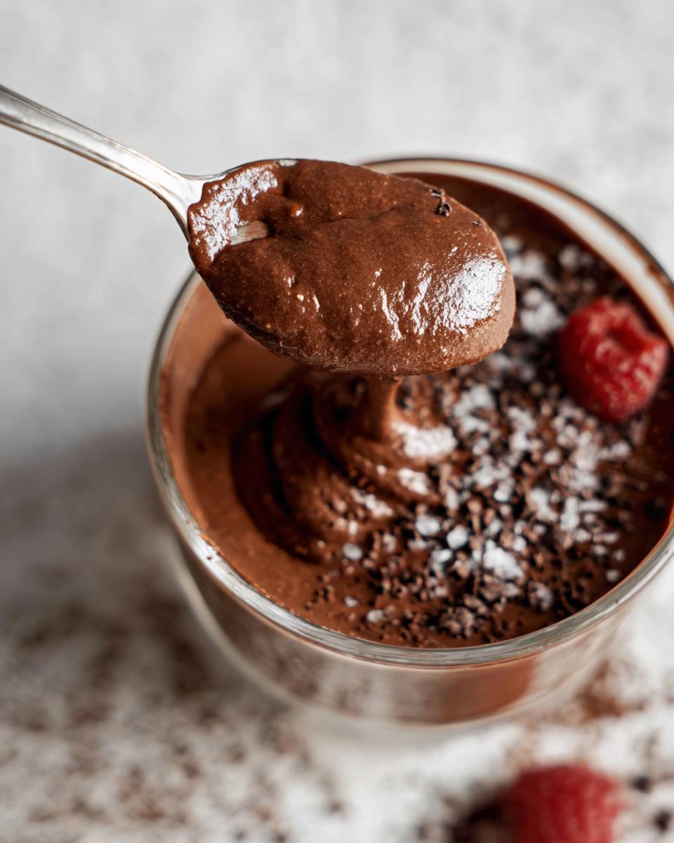 A spoonful of rich, creamy High Protein Chocolate Pudding, garnished with cocoa powder, chocolate shavings, and a raspberry.