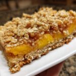 A close-up of a peach crumble bar with a buttery oat topping, showcasing the layers of fruit and crust.